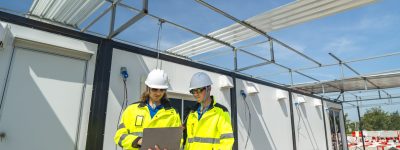 Two engineers in high visibility jackets and helmets working on a laptop at a modular construction site, with steel framework and wiring visible, highlighting teamwork, planning construction.